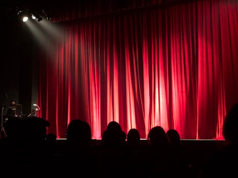 pexels-photo-713149-713149 Dimly lit theater stage with red curtains and audience silhouettes under spotlights.